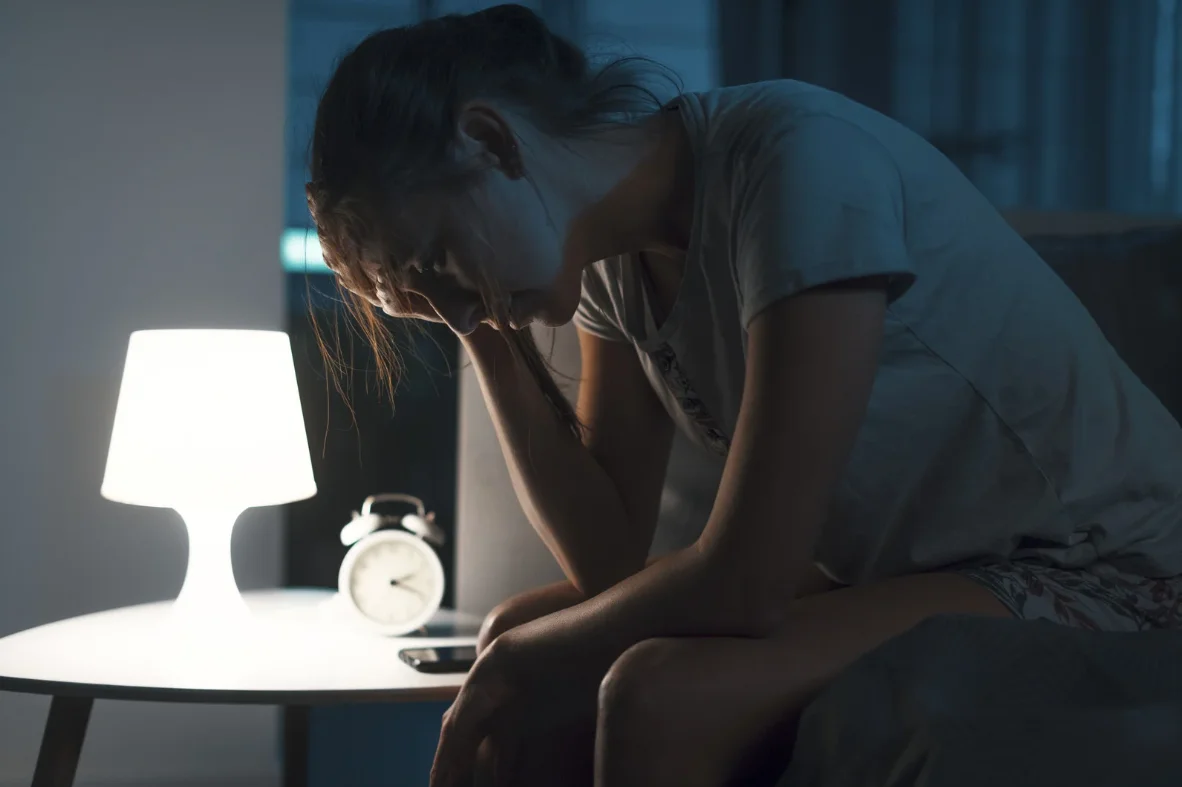 Chica sentada en la cama con la mano en la cabeza, luz de mesita de noche encendida y reloj, mostrando síntomas de ansiedad nocturna e insomnio.